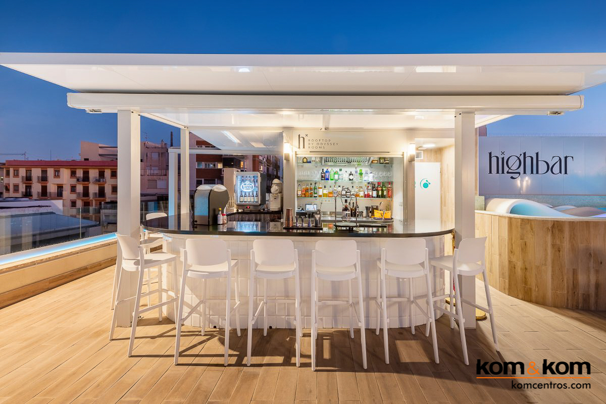 Bar de terraza en azotea de hotel con encimera de acero inoxidable, estantes de cristal con botellas de c&oacute;ctel variadas, cocteleras y fregadero. Dise&ntilde;o elegante y profesional para bar de exteriores.
